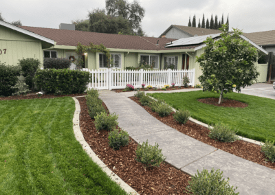 A single-story house with light green siding, a brown roof, white fence, landscaped yard, and a concrete walkway curving through mulch and bushes.