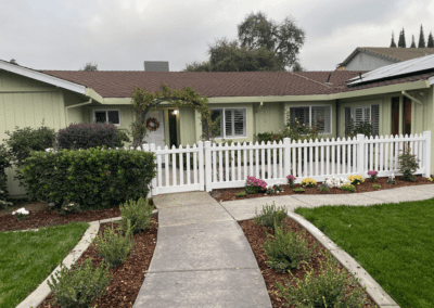 Single-story house with light green siding, brown roof, white picket fence, landscaped yard, and a curved concrete path leading to the front door.