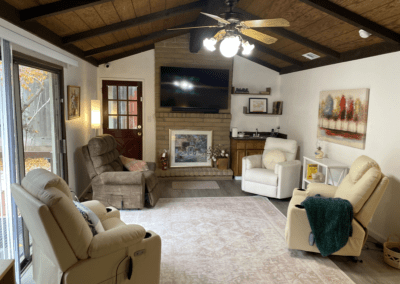 A cozy living room with three recliners, a TV mounted above a fireplace, a ceiling fan, wall art, and sliding glass doors leading outside.