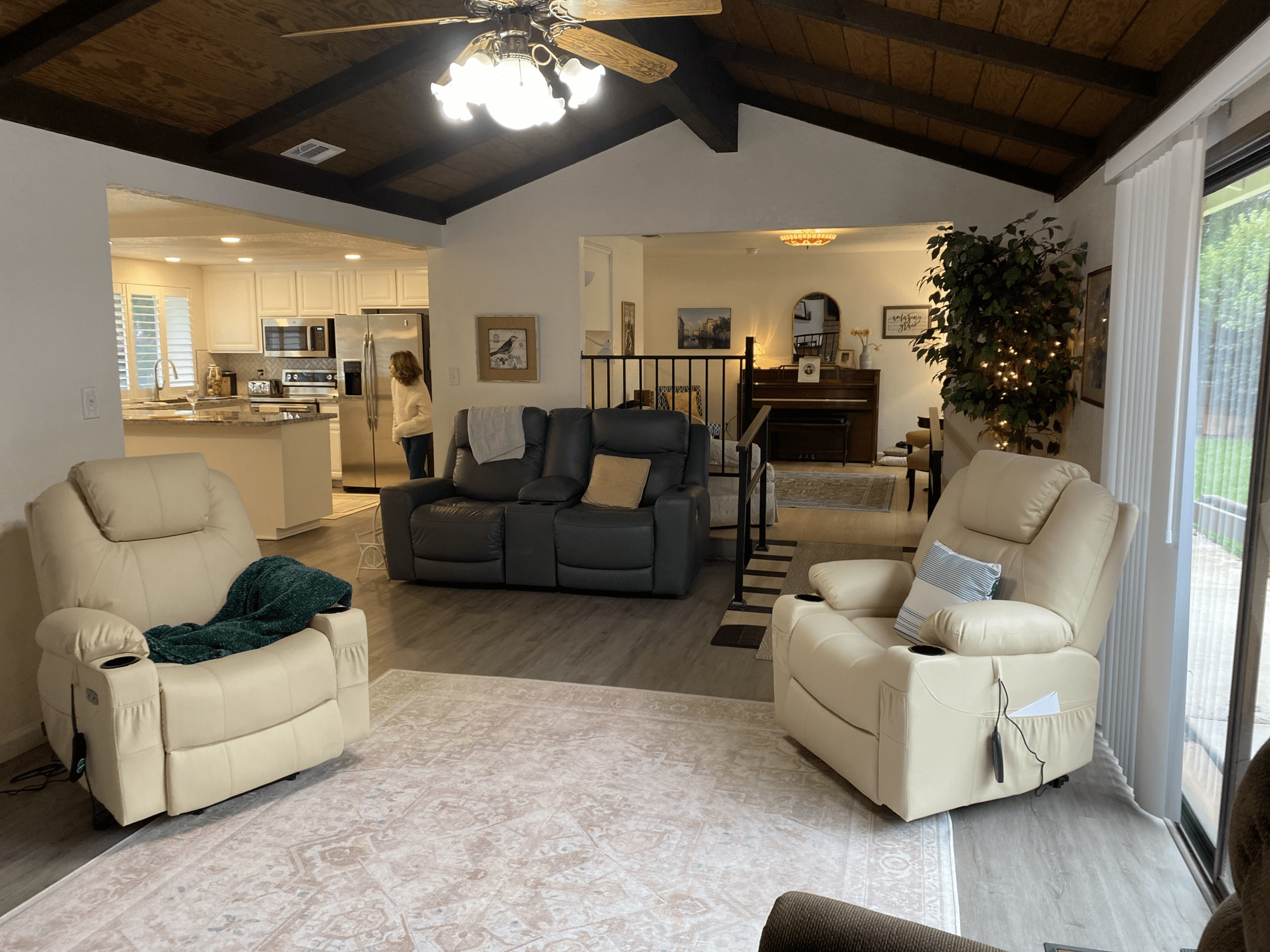 A spacious living room with beige recliners, a dark loveseat, a piano in the dining area, and a person walking in the kitchen. Ceiling fan and large windows provide natural light.