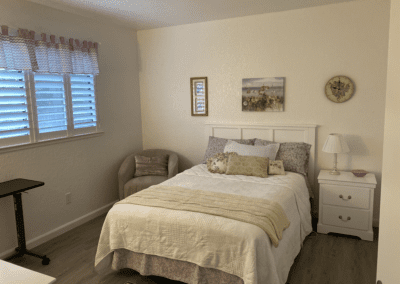 A neatly made bed with pillows and a throw blanket is next to a nightstand, lamp, and armchair in a softly lit bedroom with wall art and shuttered windows.