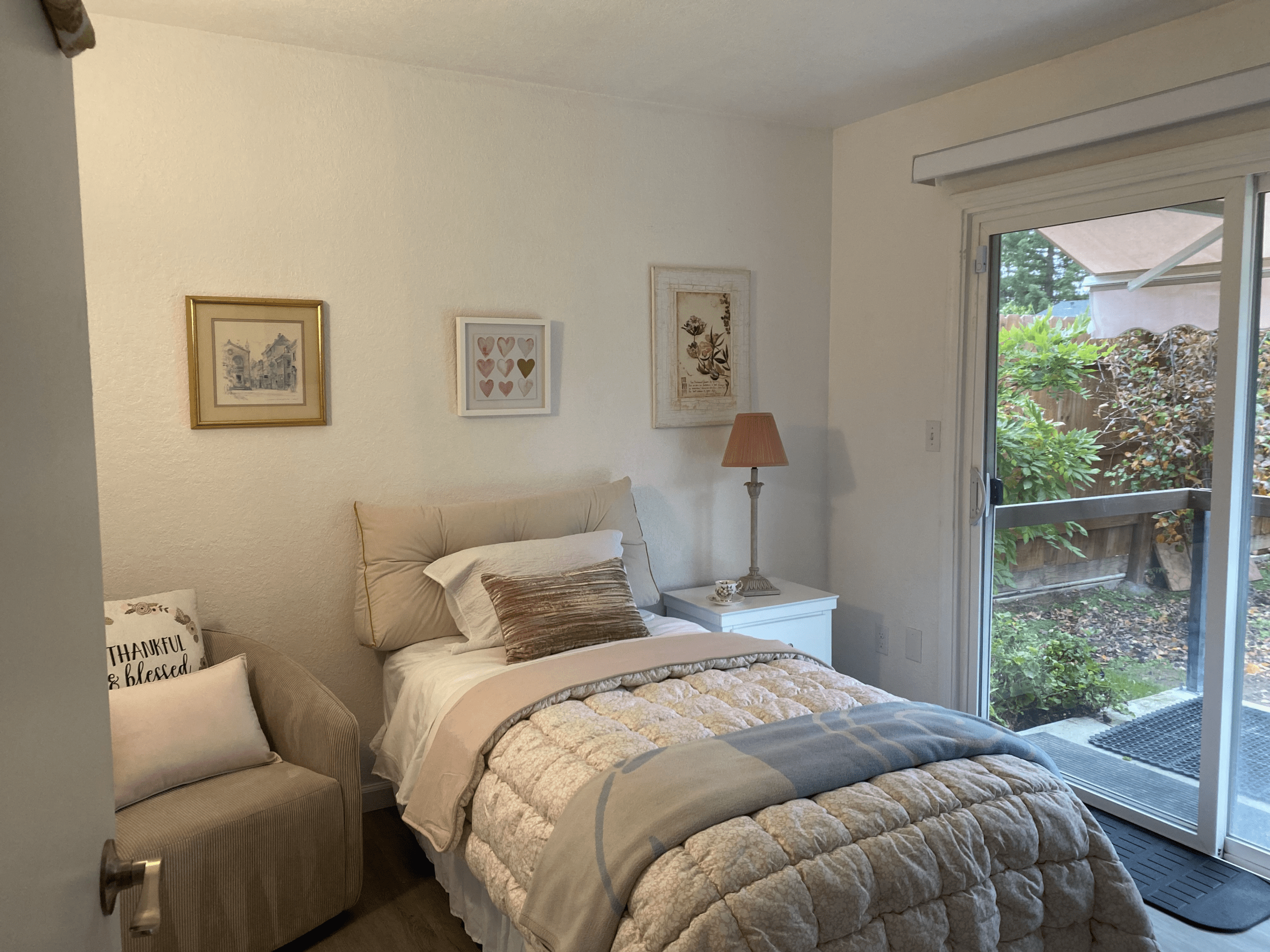 A small, neatly made bedroom with a single bed, armchair, framed art on the wall, a nightstand with a lamp, and a sliding glass door leading to a garden.
